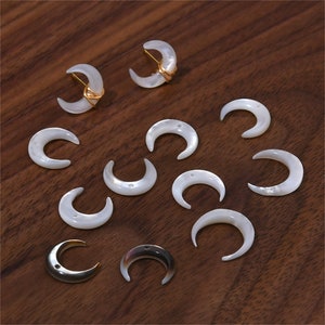 Op de afbeelding: Een verzameling witte halve maanvormige schelpbedels met twee gouden draadgewikkelde bedels. De bedels liggen op een houten oppervlak.