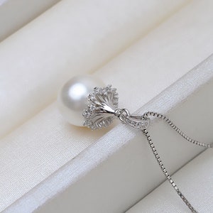 Puede incluir: Un collar de plata con un gran colgante de perla blanca. El colgante tiene un diseño decorativo de corona de plata con pequeñas piedras transparentes.