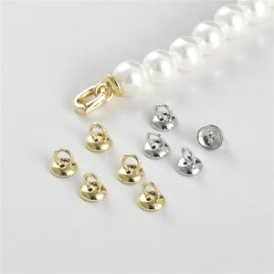 Puede incluir: Primer plano de cierres de metal dorado y plateado para la elaboración de joyas. Los cierres son redondos con un pequeño bucle en la parte superior. Hay seis cierres dorados y tres cierres plateados.