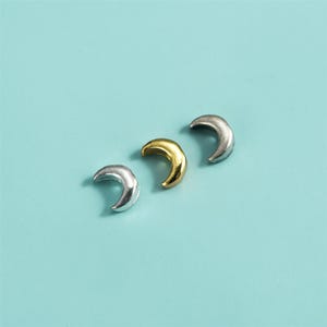 May include: Three crescent moon charms in silver and gold tones are arranged on a light blue surface. The charms are small, with a curved shape, and appear to be made of metal. The gold charm is in the center.