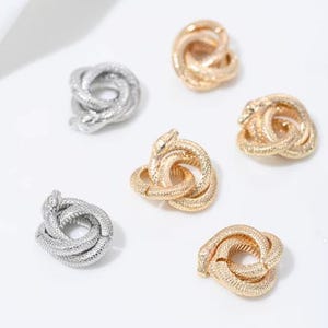 May include: An assortment of gold and silver-toned snake-shaped jewelry pieces. The jewelry is arranged on a white surface. The snakes are intertwined, creating a knot-like design. The jewelry has a textured surface.