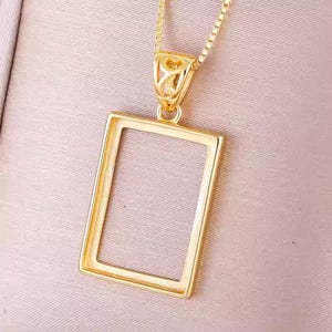 Puede incluir: Un collar con un colgante rectangular de color dorado. El marco está abierto y listo para contener una foto u otro objeto pequeño.