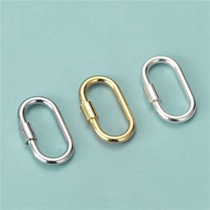 May include: Three oval-shaped carabiner clasps in silver and gold tones. The clasps are shown against a light teal background. The center clasp is gold, while the other two are silver. These clasps are often used for jewelry making or crafting.