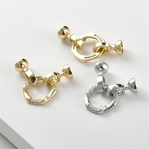 Puede incluir: Tres cierres de joyería metálicos en tonos dorados y plateados. Cada cierre presenta un anillo decorativo con pequeñas piedras transparentes y dos extremos redondeados. Los cierres están diseñados para la fabricación de joyas y manualidades.