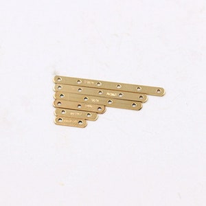 Peut inclure: Un ensemble de huit sangles métalliques dorées avec des trous pour la fixation à d'autres objets. Les sangles sont disposées en éventail, la plus grande en haut et la plus petite en bas. Les sangles ont toutes la même largeur, mais des longueurs différentes. Chaque sangle a un nombre différent de trous, allant de quatre à huit.