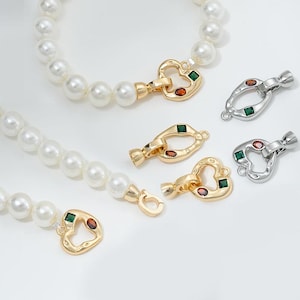 May include: Two pearl necklaces with gold and silver heart-shaped clasps with green and red gemstones. The clasps are in different styles.