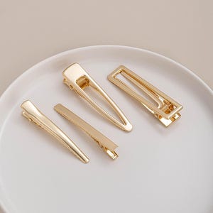 May include: Four gold-coloured hair clips of varying shapes and sizes are arranged on a white plate. The clips include a rectangular barrette, a triangular clip, and two smaller, solid clips. The clips have a shiny, metallic finish.