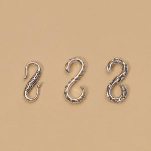 May include: Three silver-toned metal S-hooks arranged in a row. The first hook features a smooth, banded design. The second hook has a textured, ridged surface. The third hook displays a rugged, uneven, and hammered-style finish on its surface.