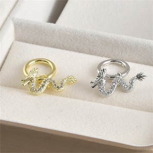 Può includere: Due orecchini a cerchio con un ciondolo a forma di drago. Il drago è tempestato di strass trasparenti. Un orecchino è dorato e l'altro è argentato.