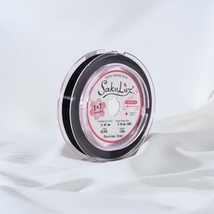 May include: A spool of black nylon-coated wire, labeled "SakuLux", with a pink and white label. The label indicates a 0.40 mm coated outer diameter and a 0.24 mm inner rope diameter. The wire is 100 meters long and made of stainless steel.