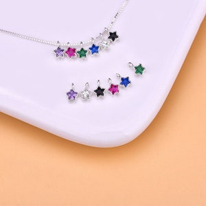 May include: A set of silver star-shaped charms with colorful gemstones. The charms are arranged in a row on a white surface. The charms are perfect for adding a touch of sparkle to jewelry.