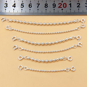 May include: Several silver ball chain extenders with lobster claw clasps and jump rings are displayed against a light tan background. The extenders vary in length, with the longest measuring approximately 8 inches.