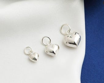 Könnte beinhalten: Drei silberne herzförmige Anhänger in verschiedenen Größen, jeweils mit einer kleinen Schlaufe oben. Sie werden auf einem weißen Stoff mit einem dunkelblauen Hintergrund präsentiert. Die Anhänger haben eine polierte, reflektierende Oberfläche.