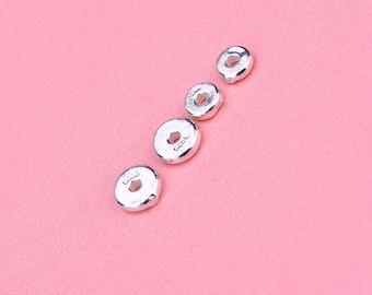 May include: Four small, silver-colored, disc-shaped beads with a hole in the center are arranged diagonally on a pink background. The beads have a slightly textured surface and a shiny finish. The beads are likely for jewelry making.