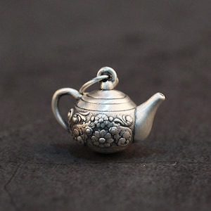 May include: A small, silver-toned metal charm shaped like a miniature teapot. The body of the teapot features a detailed, raised floral relief pattern, a curved handle, a short spout, and a small circular loop at the top for attachment.