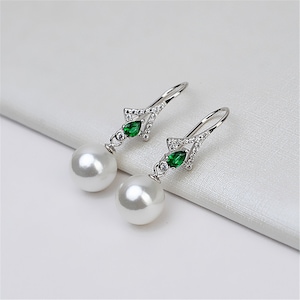 Puede incluir: Un par de pendientes de plata con una perla blanca y una piedra preciosa verde. Los pendientes tienen un diseño delicado con pequeñas piedras transparentes.