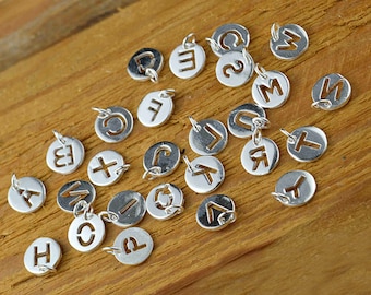 May include: A collection of silver charms featuring various letters and symbols. The charms are round and have a hole at the top for attaching to a necklace or bracelet.