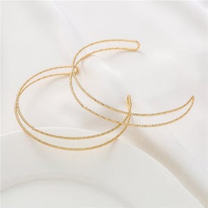 May include: A gold-coloured headpiece with a crescent moon design. The headpiece is made of thin, textured metal and is displayed on a white surface. The design features multiple curved lines, creating an elegant and minimalist aesthetic.