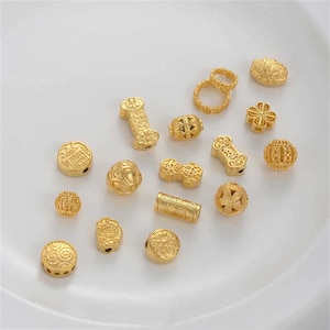 Könnte beinhalten: Eine Sammlung goldfarbener Perlen und Anhänger in verschiedenen Formen, darunter runde, zylindrische und florale Designs. Einige Perlen weisen aufwendige Muster und Ausschnitte auf. Die Artikel werden auf einer weißen Oberfläche präsentiert.