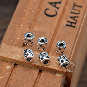 May include: Seven silver metal beads with a star design. The beads are round and have a hole in the center. They are arranged in a row on a wooden surface.