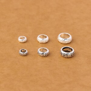 Peut inclure: Six perles en métal argenté disposées en deux rangées de trois, chacune présentant un motif de volutes en relief répété. Les perles varient en taille, de la plus petite à gauche à la plus grande à droite, avec une finition métallique.