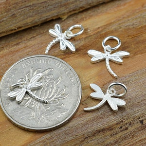 May include: Four silver dragonfly charms with a loop for attaching to jewelry. The charms are on a wooden surface.