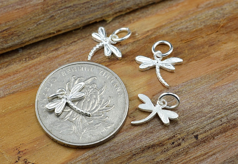 May include: Four silver dragonfly charms with a loop for attaching to jewelry. The charms are on a wooden surface.
