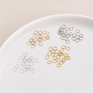 May include: A collection of small, oval-shaped jump rings in gold and silver tones. The jump rings are scattered on a white surface, with several groupings of the rings visible. These jewelry findings are ideal for crafting.