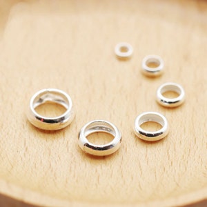 May include: A set of six silver jump rings of varying sizes. The rings are smooth and shiny, and they are arranged in a semi-circle on a wooden surface.