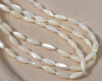 May include: Close-up of multiple strands of off-white, oval-shaped beads. The beads have a pearlescent sheen and are strung together, creating a layered effect. The background is a soft, neutral beige fabric, highlighting the beads' color and texture. The beads appear to be made of a natural material.