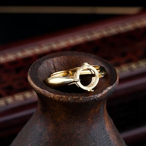 May include: A gold ring setting with an oval-shaped bezel for a gemstone. The ring is on a wooden surface.