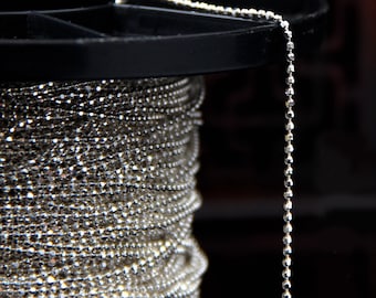 May include: A close-up of a silver chain with a 1.2 mm thickness. The chain is wound around a spool and a section of the chain hangs down. The chain is made of small, round beads.