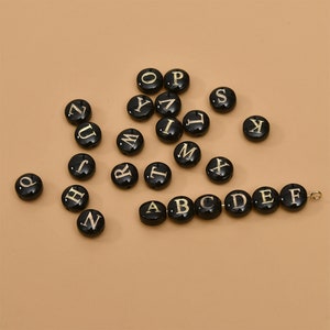 Könnte beinhalten: Schwarze Alphabet-Buchstabenperlen mit goldener Beschriftung. Die Perlen sind auf einer beigefarbenen Oberfläche verstreut. Eine Perlenkette mit den Buchstaben A bis F ist aufgereiht.