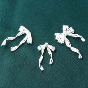 Pode incluir: Três pingentes de prata em forma de laço com um design de fita torcida. Os pingentes são perfeitos para adicionar um toque de elegância a joias ou artesanato.