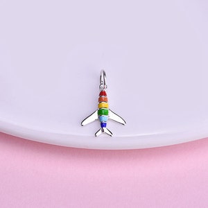 Könnte beinhalten: Ein silberner Flugzeug-Anhänger mit einem Regenbogen-Emaille-Design. Der Anhänger ist perfekt, um jedem Schmuckstück einen Hauch von Verspieltheit zu verleihen.