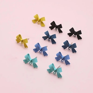 Może przedstawiać: Kolekcja kolczyków sztyftów w kształcie kokardek w różnych kolorach, w tym żółtym, czarnym, turkusowym i odcieniach niebieskiego. Kolczyki ułożone są na jasnoróżowej powierzchni, prezentując ich delikatny design i żywe kolory.