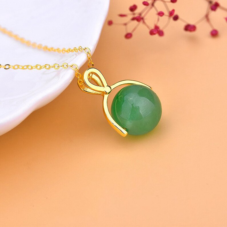 May include: A gold-colored necklace with a green jade sphere pendant. The pendant is held in place by a gold-colored bow-shaped frame. The chain is delicate and gold-colored. The background is a soft orange.