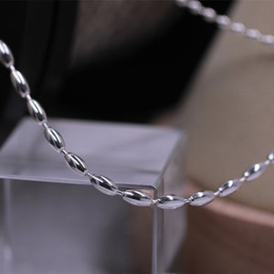 Peut inclure: Un collier en chaîne argent avec des maillons de forme ovale. La chaîne est posée sur une surface en verre transparent.