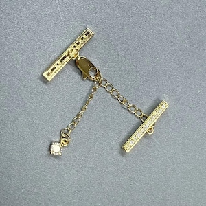 Puede incluir: Un cierre de collar dorado con un diseño de barra en T. Una barra en T está adornada con pequeños cristales transparentes. Una delicada cadena con un solo dije de cristal cuadrado cuelga del cierre. El cierre está sobre un fondo gris.