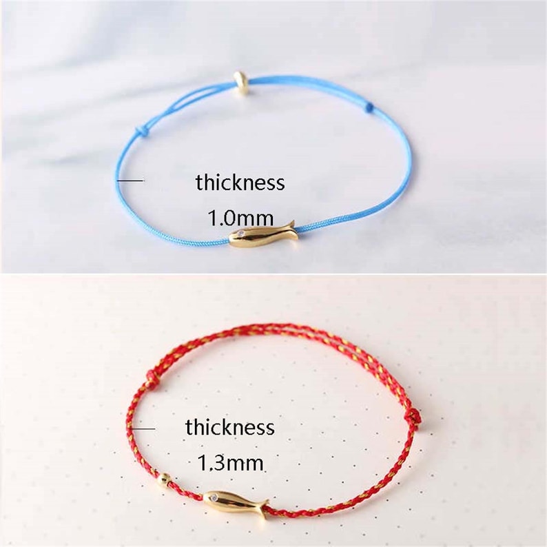 May include: Two bracelets with a gold fish charm. The top bracelet is light blue with a 1.0mm thickness. The bottom bracelet is red with a 1.3mm thickness.