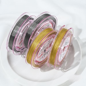May include: Spools of wire in various colors, including silver and gold, are displayed on a white plate. The wire is wound around clear plastic spools with pink edges. The spools are labeled with text, and the wire appears to be for jewelry making.
