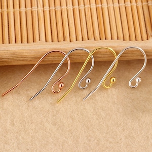 Puede incluir: Cuatro ganchos de pendientes metálicos en cobre, plata, oro y plata. Cada gancho tiene un diseño curvo con una pequeña bola en la parte inferior. Los ganchos están dispuestos sobre una superficie marrón claro.