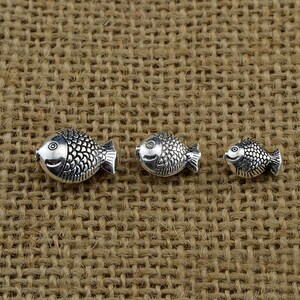 May include: Three silver fish charms of varying sizes. The charms are detailed with scales and have a slightly raised eye. The charms are arranged in a row on a brown burlap background.