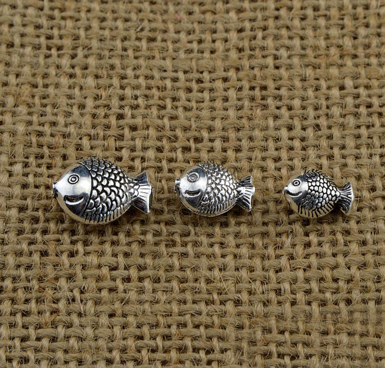 May include: Three silver fish charms of varying sizes. The charms are detailed with scales and have a slightly raised eye. The charms are arranged in a row on a brown burlap background.
