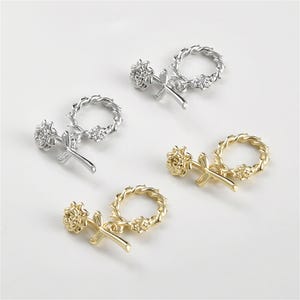 May include: Four decorative charms in silver and gold. Each charm features a flower, a cross, and a circular wreath design. The charms are arranged on a white surface, with two silver and two gold charms.