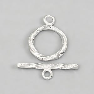 Può includere: Un set di chiusura a toggle color argento, con un design a torsione. Il set include un anello circolare con un'asola e una barra con un'asola. La chiusura è progettata per la creazione di gioielli.
