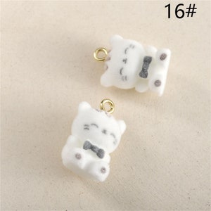 May include: Two white fuzzy cat charms with black bow ties and gold jump rings. The charms are sitting on a white surface. 16#
