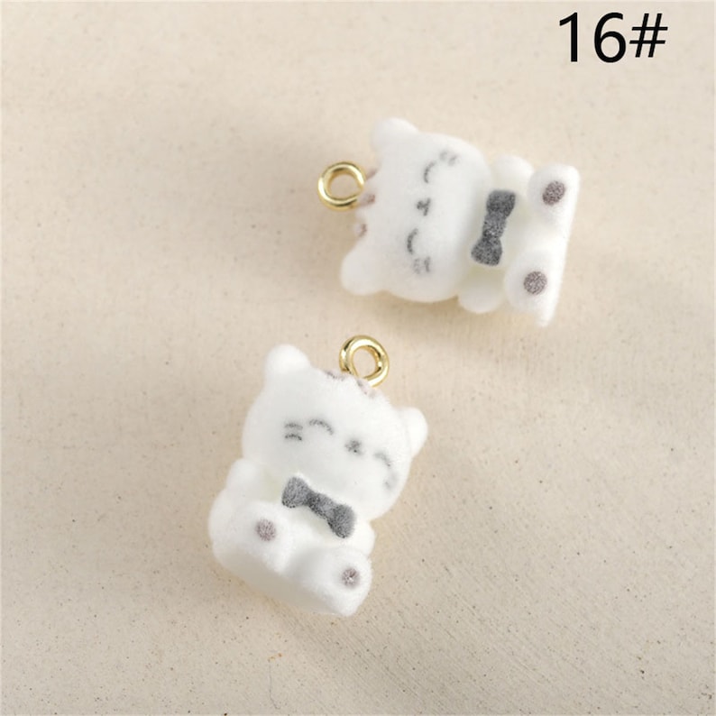May include: Two white fuzzy cat charms with black bow ties and gold jump rings. The charms are sitting on a white surface. 16#