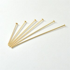 Puede incluir: Un conjunto de seis alfileres de cabeza plana de color dorado. Los alfileres est&aacute;n dispuestos en forma de abanico, con las cabezas apuntando hacia la parte superior de la imagen.