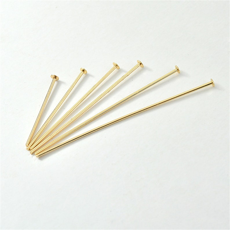 Puede incluir: Un conjunto de seis alfileres de cabeza plana de color dorado. Los alfileres est&aacute;n dispuestos en forma de abanico, con las cabezas apuntando hacia la parte superior de la imagen.